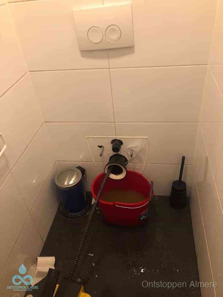 Een ontstopte wand-wc met verwijderde toiletpot waar een ontstoppingsveer in de wanduitgang verdwijnt, het achterhalen waar de verstopping zit in Almere