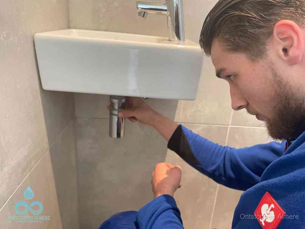 Een monteur draait met de hand de zwanenhalssifon onder een fonteinwasbak in de toiletruimte los om de bron van rioollucht in Almere te vinden
