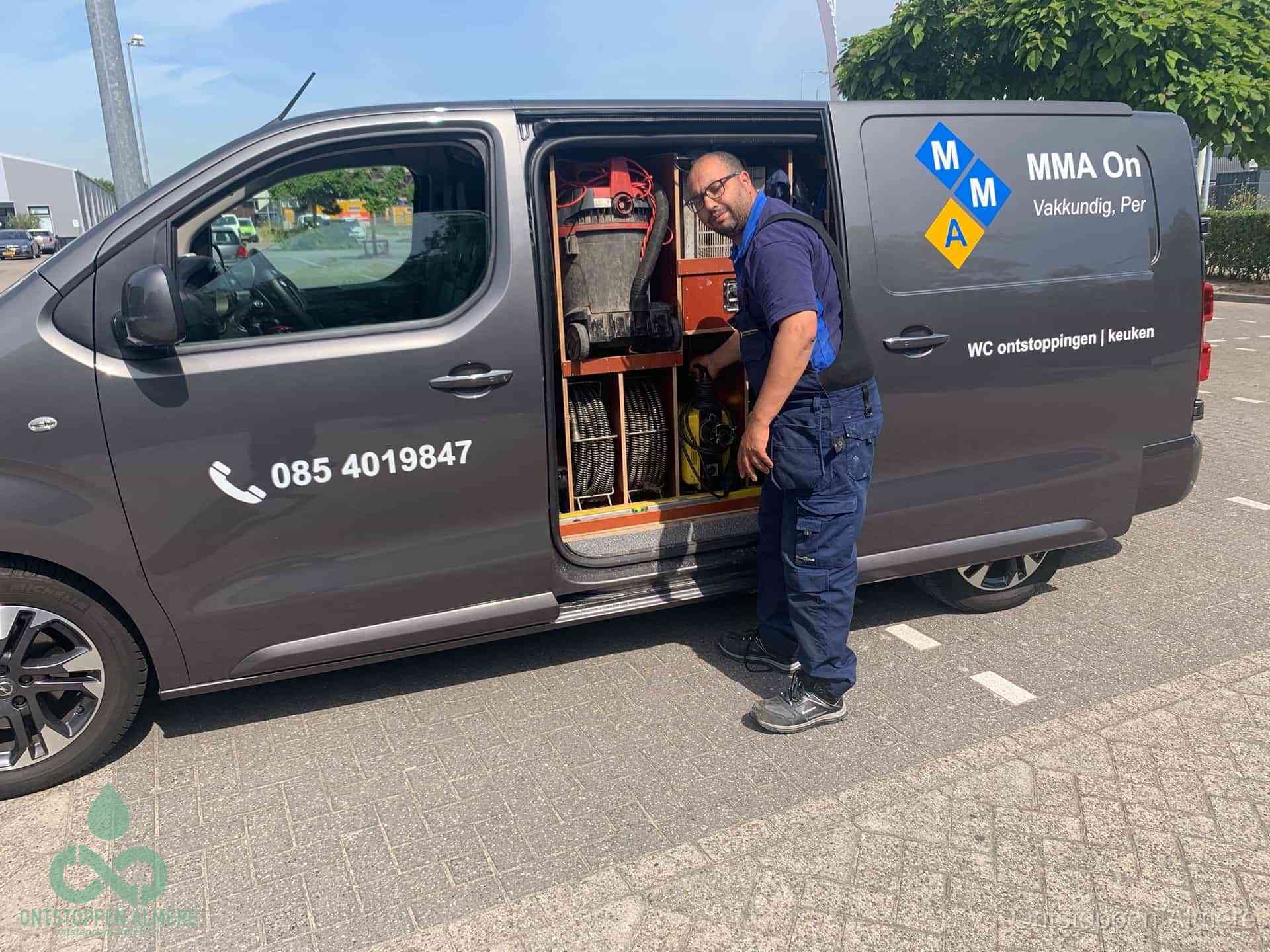 Een monteur bij de open schuifdeur van zijn bedrijfsbus op een parkeerplaats, klaar om de oorzaak van terugkerende afvoerverstoppingen aan te pakken in Almere