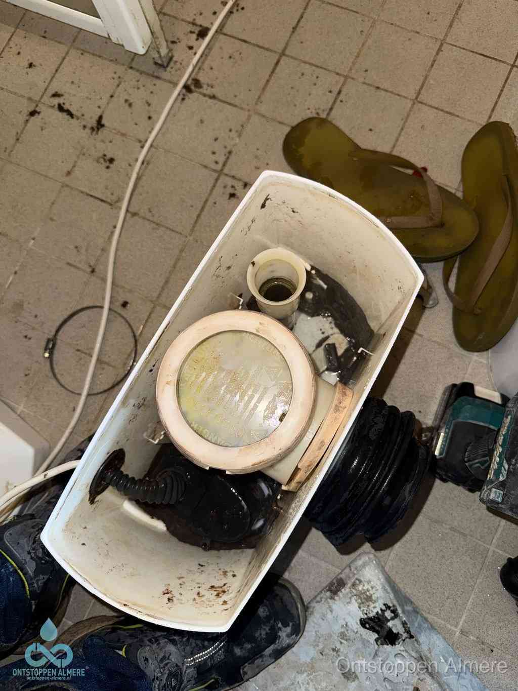 Een gedemonteerde toiletpot op de vloer met ontstoppingsveer en gereedschap ernaast, het verwijderen van een vastzittend voorwerp uit de wc in Almere